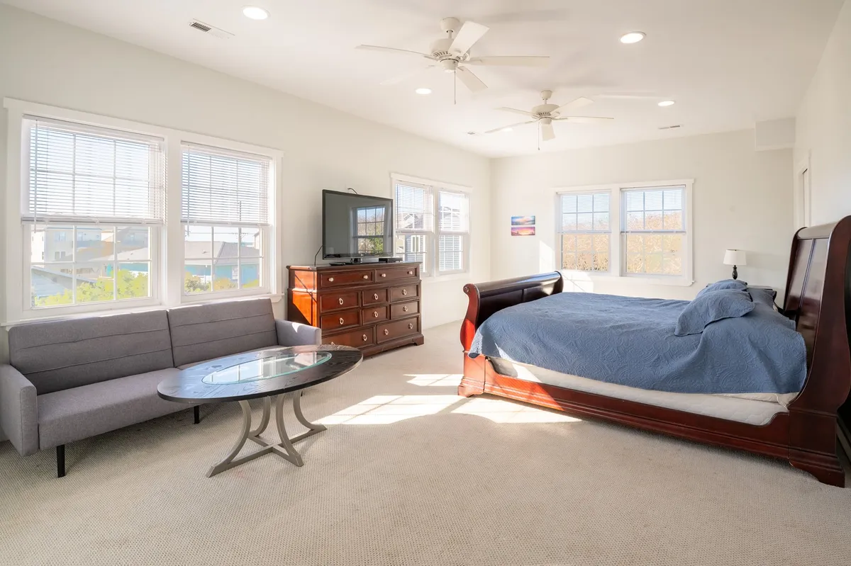 A versatile bedroom with a bed, sofa, coffee table, and a dresser, complemented by a television. Sunlight fills the space, suggesting it’s both a relaxing sleeping area and a cozy spot for socializing or watching TV.