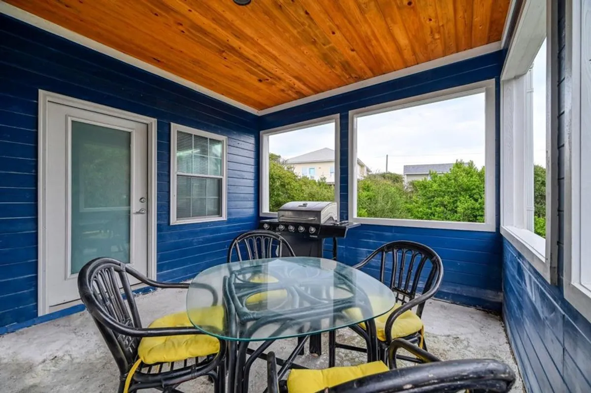 A covered porch with a glass dining table and black chairs sits under a wooden ceiling. A propane grill is ready for outdoor cooking, while lush greenery provides a private, tranquil backdrop. The seating arrangement encourages gatherings and relaxed meals.