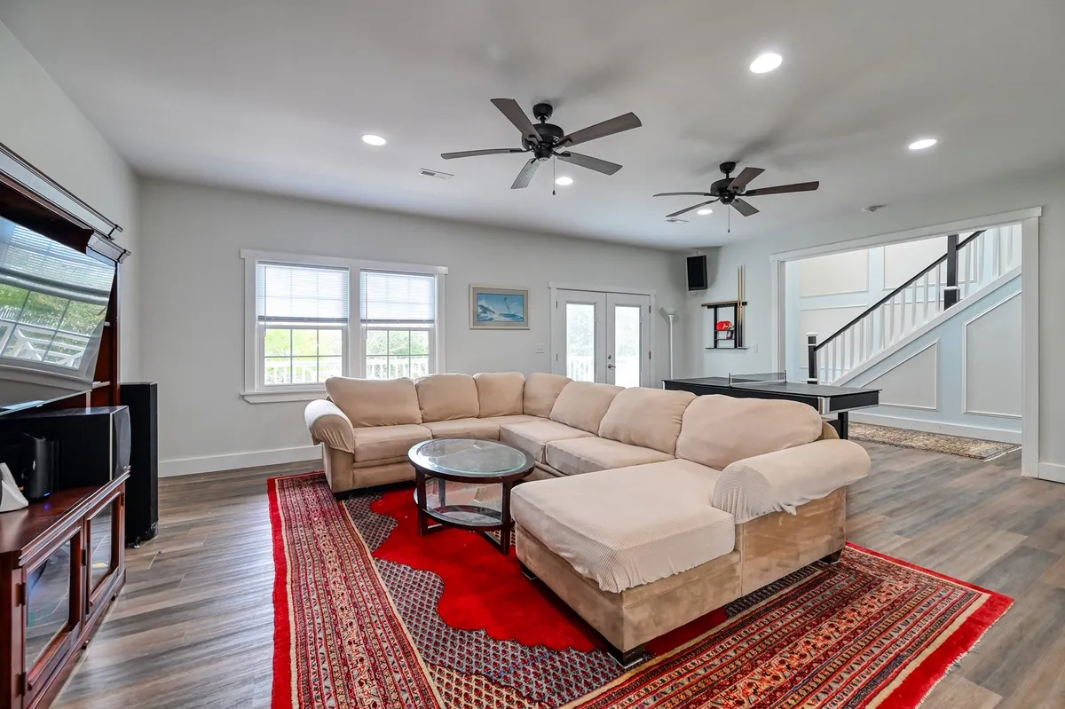 A spacious living area featuring a large sectional sofa, coffee table, rug, ceiling fans, and ample windows that flood the room with natural light. The setup invites groups to gather for movie nights or games, offering a comfortable environment for relaxation and shared activities.