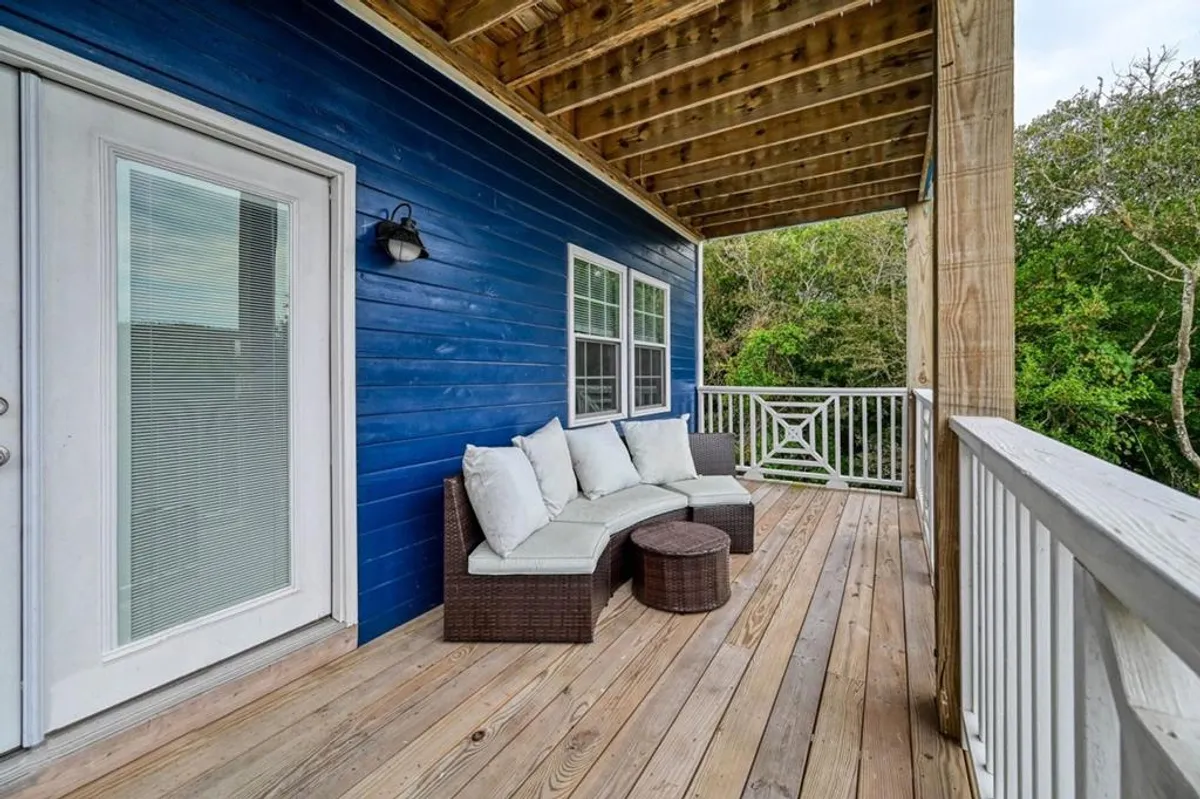 A peaceful outdoor deck area with comfortable seating, set against a blue wooden wall and surrounded by lush greenery. The setting suggests a relaxing spot to unwind after a day at the beach, with ample space to enjoy the outdoors with your pet.