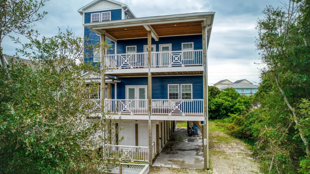 This view highlights the blue exterior of the house, its balconies with railings, and surrounding greenery. The scene conveys a welcoming, tranquil mood and hints at spacious outdoor living areas ideal for family gatherings or unwinding after a day at the beach.