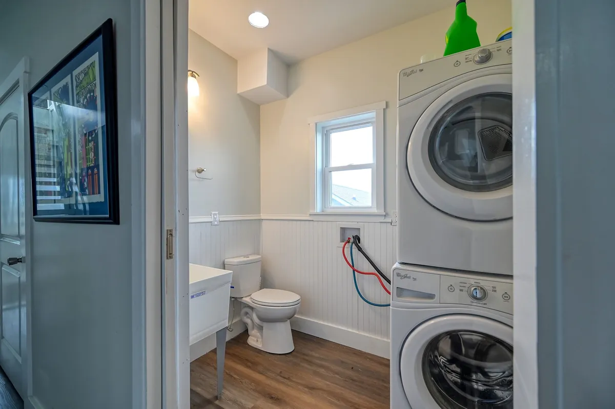 A practical, bright laundry area featuring stacked washer and dryer units, a small window letting in natural light, and a toilet. The space is tidy and purpose-driven.