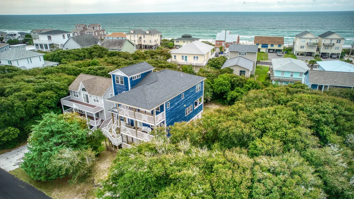 A wide shot of the blue house surrounded by greenery, with the ocean visible in the background. The perspective captures both the home and its natural surroundings, emphasizing its serene and private location near the beach.