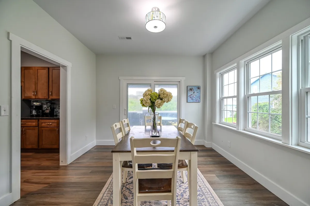 A bright, welcoming dining area centered around a large table with a floral centerpiece. Large windows bathe the space in light, while the kitchen entrance in the background shows the natural flow of the open-concept layout.