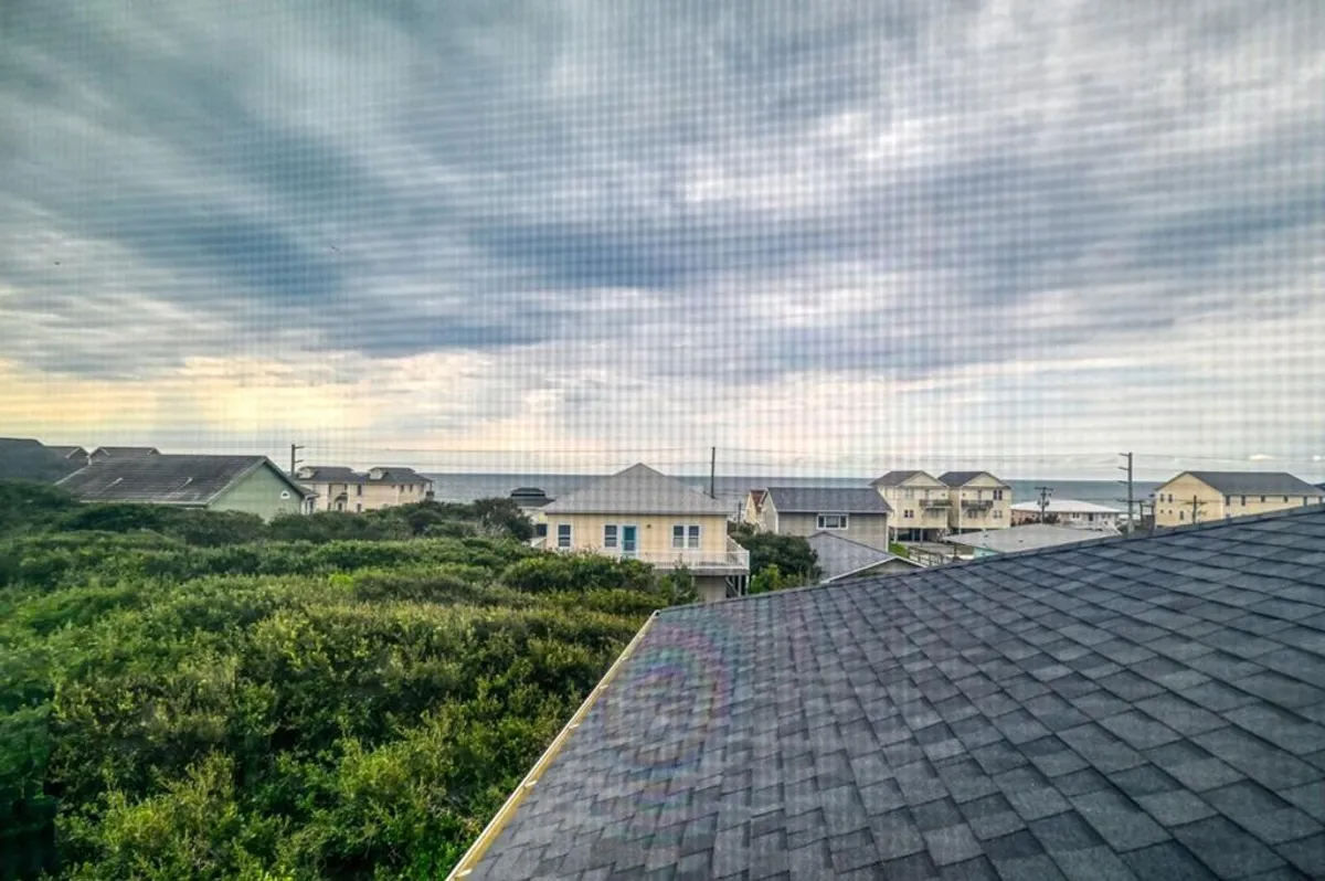 A scenic view of the ocean with a cloudy sky above, rooftops in the foreground, and greenery scattered throughout. The image evokes the feeling of being close to the beach and enjoying the coastal environment.