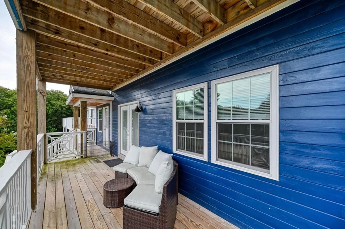 A cozy porch space with blue wooden siding, a comfortable seating arrangement, and natural greenery visible in the background. The intimate area invites lounging, sipping drinks, or reading outdoors in comfort and style.